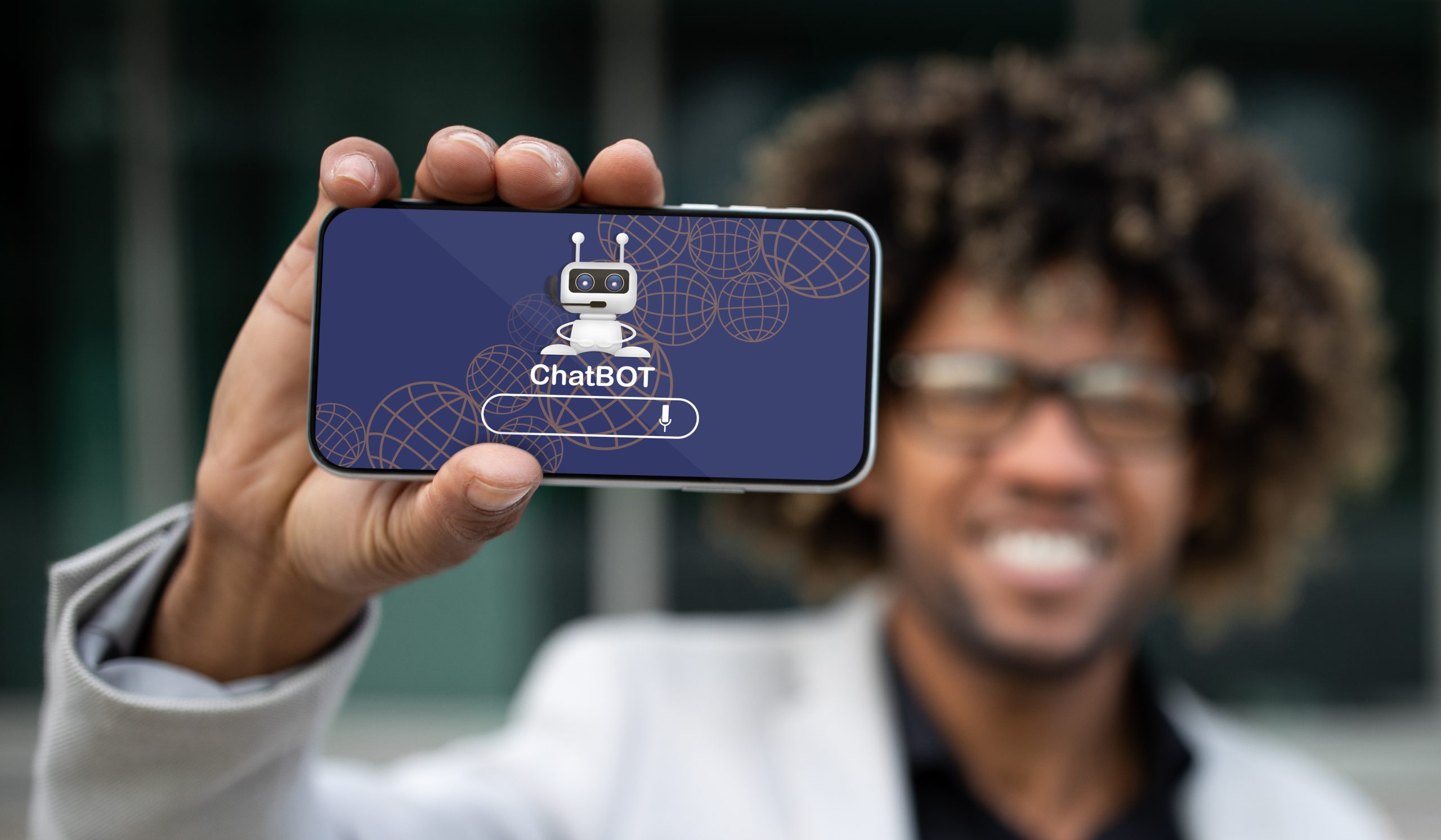 Im Hintergrund unscharfer aber sehr freundlich wirkender afroamerikanischer Unternehmer zeigt Smartphone mit Chatbot-Roboter und leerem Fragefeld